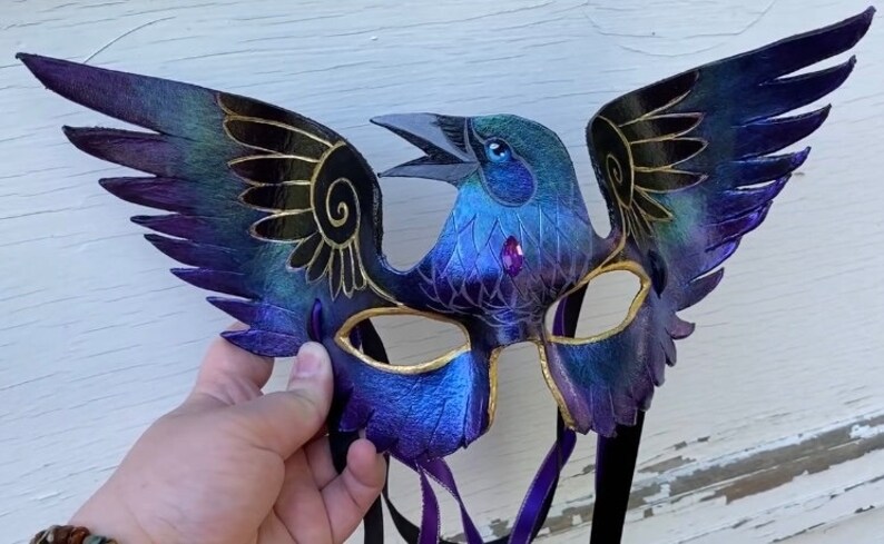 May include: A purple and blue leather masquerade mask with gold accents and a bird design. The mask has two wings and a bird's head with a jeweled eye.
