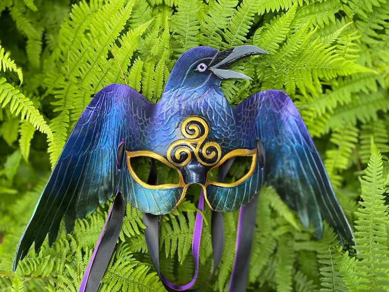 May include: A blue and purple raven mask with gold accents and a triple spiral design. The mask has black and purple ribbon ties.