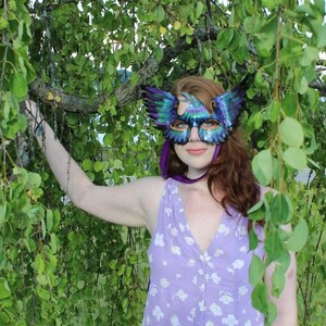 May include: A woman wearing a purple floral dress and a blue and green masquerade mask with feathers. The mask has a bird design. She is standing in front of a green leafy background.