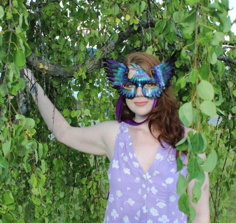 May include: A woman wearing a purple floral dress and a blue and green masquerade mask with feathers. The mask has a bird design. She is standing in front of a green leafy background.