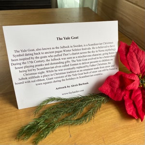 May include: A close-up of a card with text about the Yule Goat, a Scandinavian Christmas tradition. The card is on a wooden surface with a red poinsettia and green pine branches.