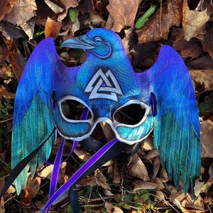 May include: A blue and green raven mask with a silver Valknut symbol. The mask has a purple ribbon attached to it.