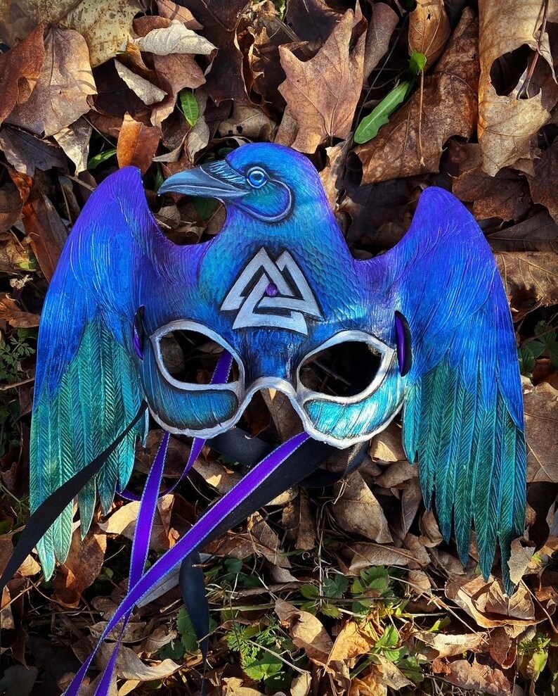 May include: A blue and green raven mask with a silver Valknut symbol. The mask has a purple ribbon attached to it.