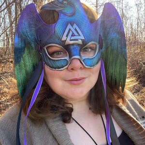 May include: A purple, green, and silver masquerade mask with a raven design and a silver Valknut symbol. The mask has black and purple ribbons attached.