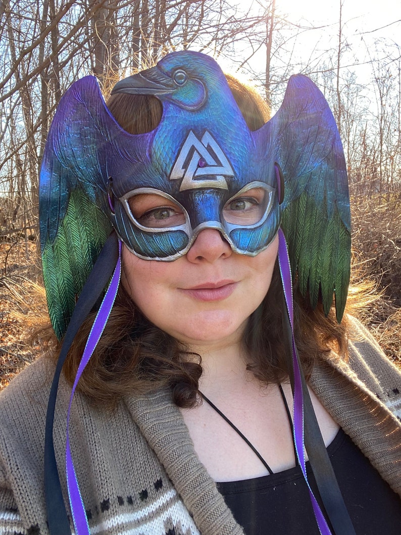 May include: A purple, green, and silver masquerade mask with a raven design and a silver Valknut symbol. The mask has black and purple ribbons attached.