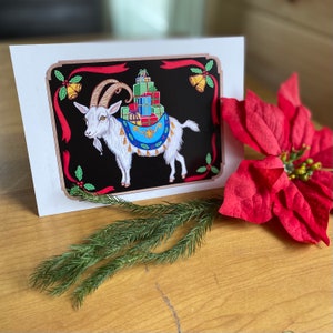 May include: A Christmas card featuring a white goat with gold horns carrying a stack of wrapped presents on its back. The goat is standing on a black background with red and green accents. The card has a festive design with holly and bells.