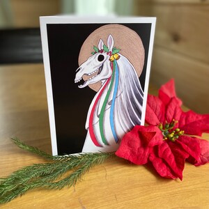 May include: A black and white illustration of a horse's skull with a white veil and colorful ribbons. The horse's skull is wearing a wreath of holly and berries. The background is black.