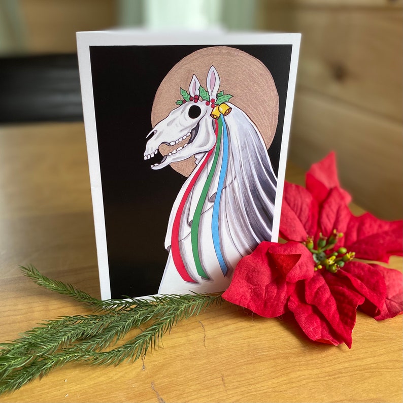 May include: A black and white illustration of a horse's skull with a white veil and colorful ribbons. The horse's skull is wearing a wreath of holly and berries. The background is black.