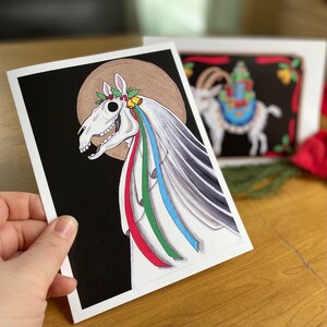 May include: A black and white illustration of a white horse skull with a wreath of holly and bells. The skull is adorned with colorful ribbons, including red, green, blue, and white.