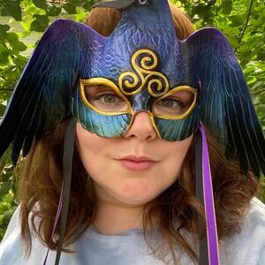 May include: A blue and purple raven masquerade mask with gold accents and a triple spiral design. The mask has black and purple ribbons attached.