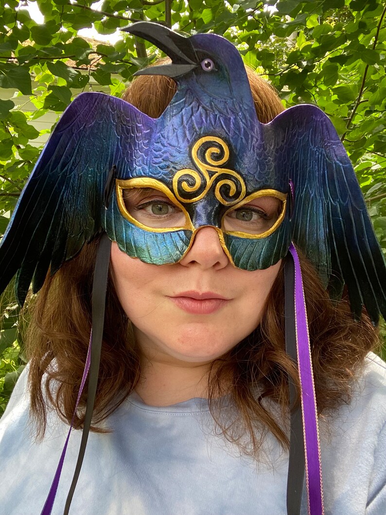 May include: A blue and purple raven masquerade mask with gold accents and a triple spiral design. The mask has black and purple ribbons attached.