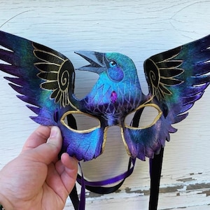 May include: A purple and blue leather raven mask with gold accents and black feathers. The mask has a detailed design and is attached to a black and purple ribbon.