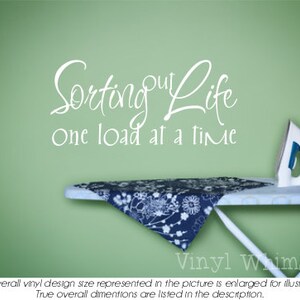 May include: White vinyl wall decal with the text "Sorting out Life One load at a time". The decal is on a green wall above an ironing board with a blue and white floral patterned ironing board cover.