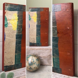 May include: Three wooden blocks painted in shades of red, brown, and green. The blocks have a distressed, vintage look and are covered in layers of paint and tape. The blocks are arranged in a row on a wooden surface. The blocks have the number "70" written on them.