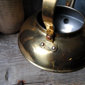 Puede incluir: Una tetera de lat&oacute;n con un asa de cobre y un peque&ntilde;o agujero en la tapa. La tetera tiene un acabado brillante y est&aacute; sobre una superficie de madera.