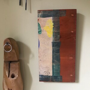 May include: A rectangular wooden panel with a layered, abstract design. The panel is painted in shades of brown, green, blue, and yellow. The number "73" is visible on the panel.