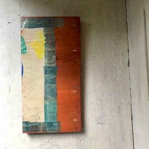 May include: A rectangular wooden panel with a layer of paper adhered to the surface. The paper has a blue, green, and yellow design with the number "73" visible. The panel is painted a reddish-brown color.