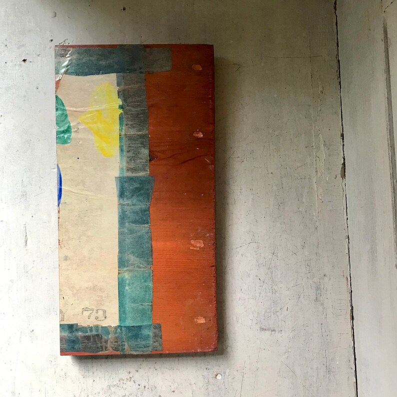 May include: A rectangular wooden panel with a layer of paper adhered to the surface. The paper has a blue, green, and yellow design with the number "73" visible. The panel is painted a reddish-brown color.
