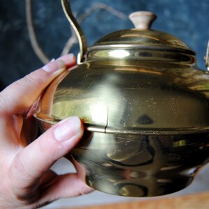 Puede incluir: Una tetera de metal de color dorado con un pomo de madera en la tapa. La tetera tiene un aspecto desgastado y envejecido con ara&ntilde;azos y una grieta en el lateral.
