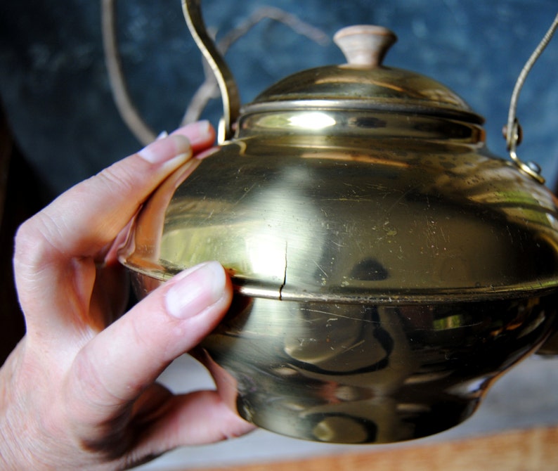 Puede incluir: Una tetera de metal de color dorado con un pomo de madera en la tapa. La tetera tiene un aspecto desgastado y envejecido con ara&ntilde;azos y una grieta en el lateral.