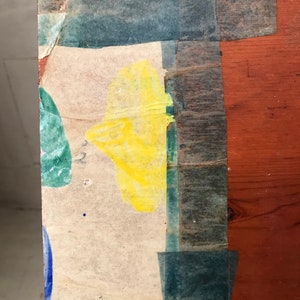 May include: A close-up of a wooden surface with a piece of paper taped to it. The paper has a yellow, green, and blue paint splatter design. The paper is taped to the wood with blue tape.