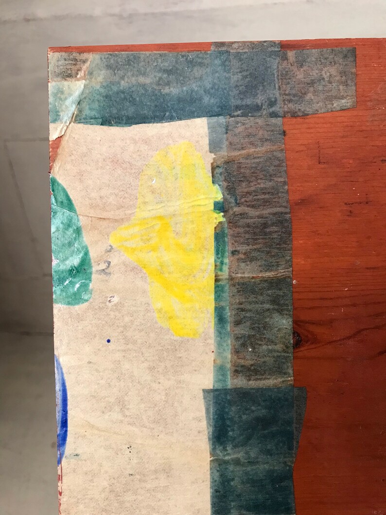May include: A close-up of a wooden surface with a piece of paper taped to it. The paper has a yellow, green, and blue paint splatter design. The paper is taped to the wood with blue tape.
