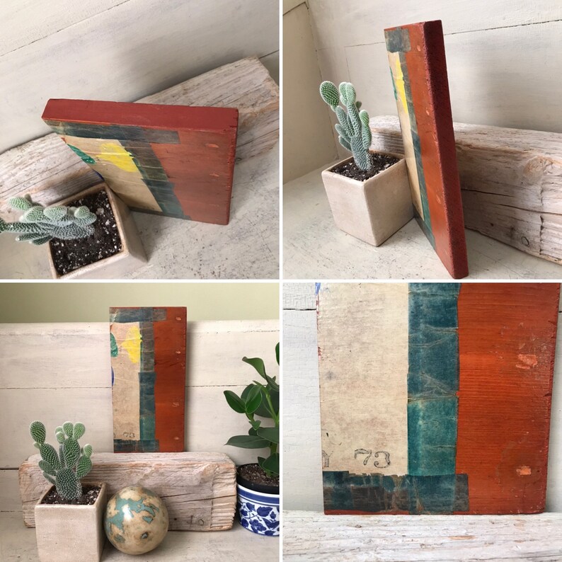 May include: A rectangular wooden sign with a layered design. The sign is painted in shades of red, yellow, green, and blue. The sign is leaning against a white wooden surface.