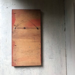 May include: A rectangular wooden sign with a light brown wood grain and a red painted border. The sign has two small metal hooks on the top for hanging.