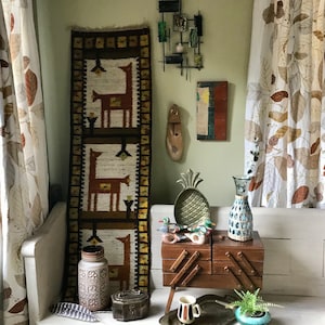 May include: A vintage wooden bench with a brown wooden box with a geometric design on top. The box is decorated with metal accents. There is a brown and white woven tapestry hanging on the wall behind the bench. The tapestry features a repeating pattern of stylized deer.