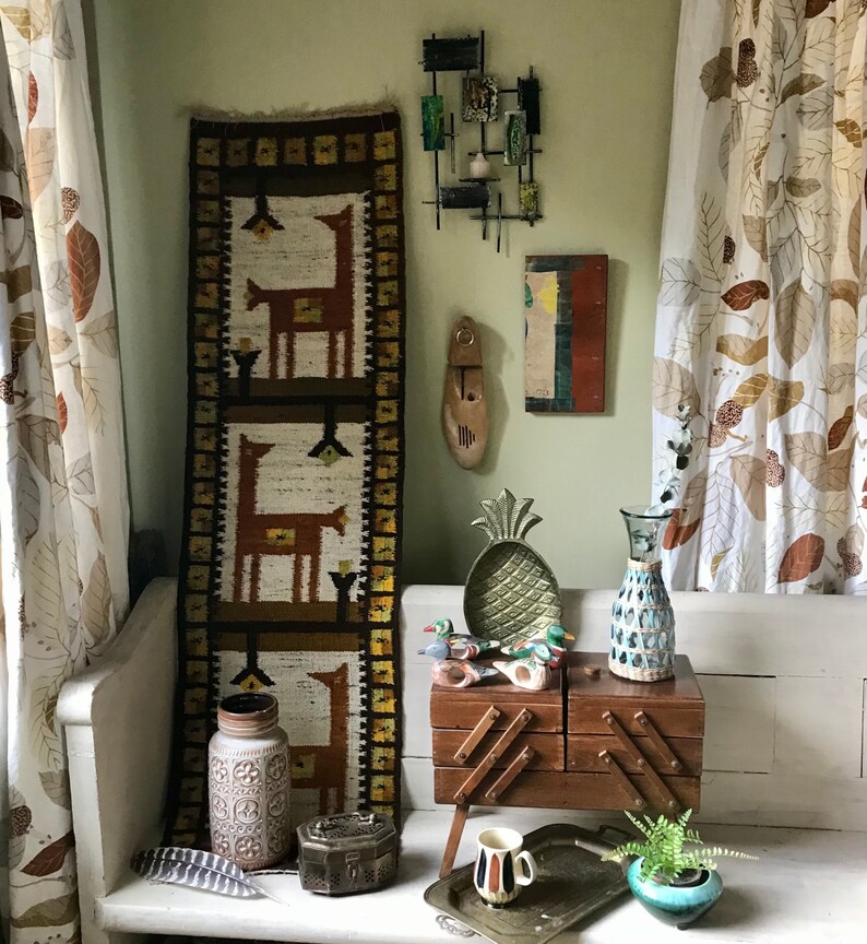 May include: A vintage wooden bench with a brown wooden box with a geometric design on top. The box is decorated with metal accents. There is a brown and white woven tapestry hanging on the wall behind the bench. The tapestry features a repeating pattern of stylized deer.