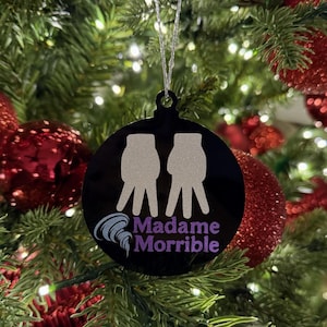 May include: Black Christmas ornament with a silver string, featuring two stylized hands and the text "Madame Morrible" in purple and white. The ornament hangs on a Christmas tree with red ornaments and lights.