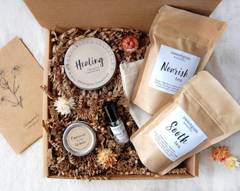 May include: A gift box filled with self-care items. The box contains two tea bags labeled "Nourish" and "Soothe", a tin of "Healing" salve, a lip balm, and a roller bottle. The box also includes a card with a chamomile illustration.