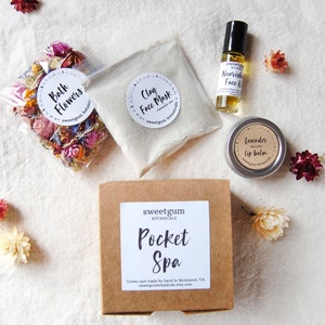 Peut inclure: Une boîte en carton marron avec le texte "sweetgum botanicals Pocket Spa" sur le devant. La boîte est entourée de divers produits de spa, dont un sac de fleurs séchées, un pot de masque facial à l'argile, un petit pot de baume à lèvres et une bouteille d'huile pour le visage.