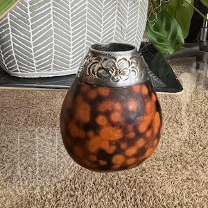 Peut inclure: Une gourde brune avec une bande argentée et un motif floral. La gourde est posée sur un tapis brun devant une plante en pot.