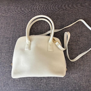 Könnte beinhalten: Cremefarbene Handtasche mit strukturierter Form, mit zwei oberen Griffen und einem abnehmbaren Schulterriemen. Die Tasche hat goldfarbene Hardware und eine glatte, matte Oberfläche. Die Tasche ist etwa 30 cm breit.