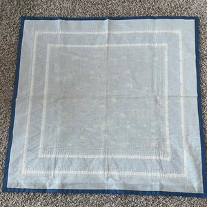以下が含まれることがあります： 白い刺繍の縁取りが施された薄い青色の正方形のテーブルクロス。テーブルクロス全体にさりげない模様があります。