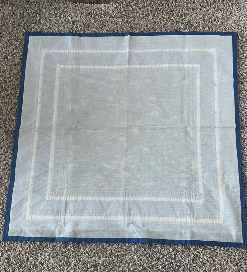 以下が含まれることがあります： 白い刺繍の縁取りが施された薄い青色の正方形のテーブルクロス。テーブルクロス全体にさりげない模様があります。