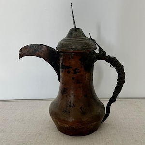 Op de afbeelding: Een antieke koperen koffiepot met een verweerde afwerking. De pot heeft een lange, gebogen tuit en een gedraaide draadhendel. De deksel is van metaal en heeft een klein, puntig knopje.