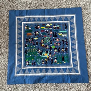 以下が含まれることがあります： 青い糸と白い糸で刺繍されたタペストリー。草原の中に人々や動物が描かれている。タペストリーは白と青の縁取りで囲まれている。