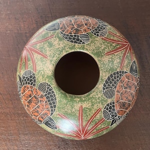 Puede incluir: Una pieza decorativa circular de cerámica con un agujero central. La pieza está pintada en tonos verdes, marrones y rojos. Tres tortugas están representadas alrededor del borde exterior, con formas rojas en forma de hoja entre ellas. Las tortugas tienen caparazones marrones y detalles negros.