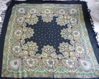 May include: A square, dark navy shawl with an intricate floral and paisley design in shades of green, gold, and cream. The center features a dark navy background with small white dots. The edges are trimmed with a fringe.