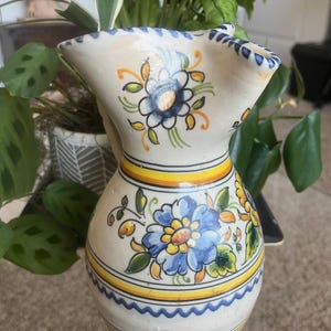 May include: A white ceramic jug with blue and yellow floral designs. The jug has a wide mouth and a handle.