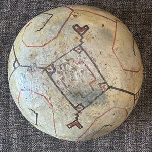 May include: A round, off-white decorative object with a geometric design. The design features red and black lines forming shapes and patterns across the surface. The object has a slightly textured appearance.