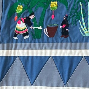 以下が含まれることがあります： 青い布製の壁掛けで、植物を収穫している人々の刺繍が施されています。シーンにはバナナの木、サトウキビの植物、そして2人の人物が描かれています。壁掛けの下部には白と青の幾何学模様が施されています。