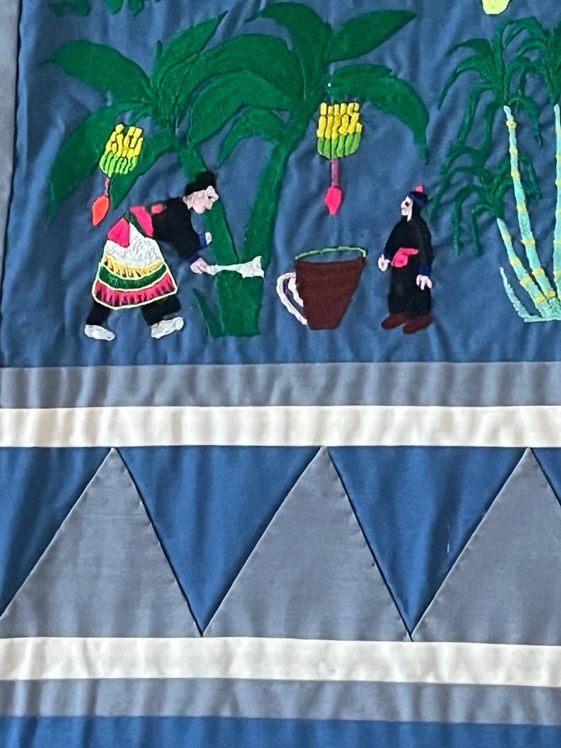 以下が含まれることがあります： 青い布製の壁掛けで、植物を収穫している人々の刺繍が施されています。シーンにはバナナの木、サトウキビの植物、そして2人の人物が描かれています。壁掛けの下部には白と青の幾何学模様が施されています。