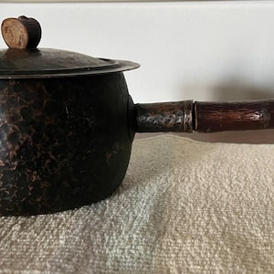 Może przedstawiać: Mały, czarny, młotkowany garnek miedziany z drewnianym uchwytem i pokrywką. Pokrywka ma mały drewniany guzik.