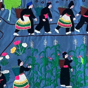 以下が含まれることがあります： 刺繍された壁掛けは、畑でトウモロコシを収穫している人々のシーンを描いています。人々は伝統的な服装をし、かごを持っています。畑は緑のトウモロコシの茎と黄色のカボチャでいっぱいです。
