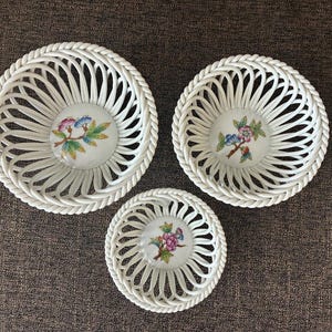 May include: Three white ceramic baskets with a woven design and floral center. The baskets are of varying sizes, with a rope-like trim around the edges. The floral centers feature pink, blue, and yellow flowers.