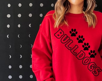 Op de afbeelding: Rood sweatshirt met zwarte tekst die "Bulldogs" zegt met poot afdrukken in een diagonale lijn.
