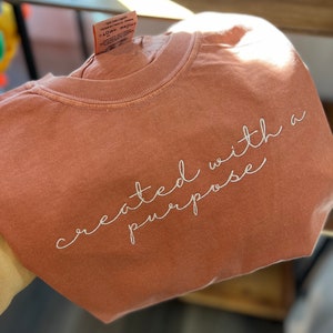 May include: A terracotta-colored t-shirt with the phrase "created with a purpose" embroidered in white script. The shirt is made of a soft material and has a classic crew neck design. A tag is visible on the collar.